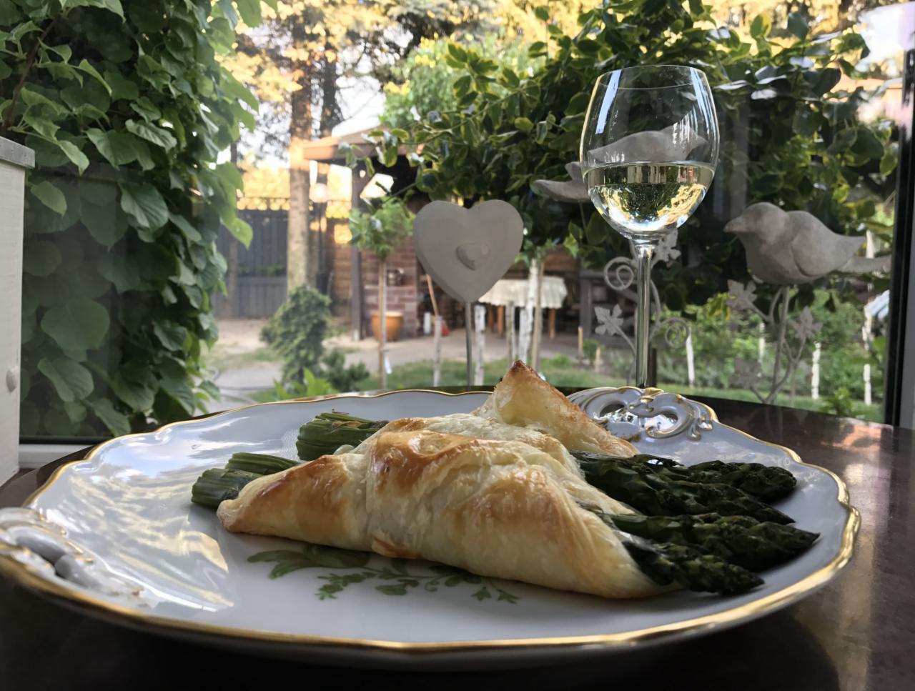 Dwa rogaliki z ciasta francuskiego wypełnione szparagami na ozdobnym talerzu z zielonymi motywami roślinnymi i złotym brzegiem, obok kieliszek białego wina, w tle ogród widoczny przez okno.