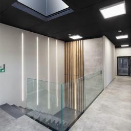 Ace Glass M. Woźnniak sp.k. - Nowoczesna klatka schodowa z szarą podłogą, szklaną balustradą z czarnymi elementami mocującymi, oświetleniem LED wzdłuż ściany i drewnianą dekoracją ścienną, widok z góry na schody.