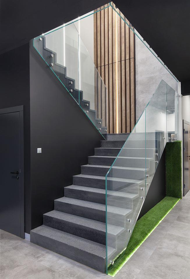 Nowoczesne schody z szarymi stopniami i szklaną balustradą mocowaną punktowo, oświetlenie LED wzdłuż drewnianej ściany i sztuczna trawa przy schodach.