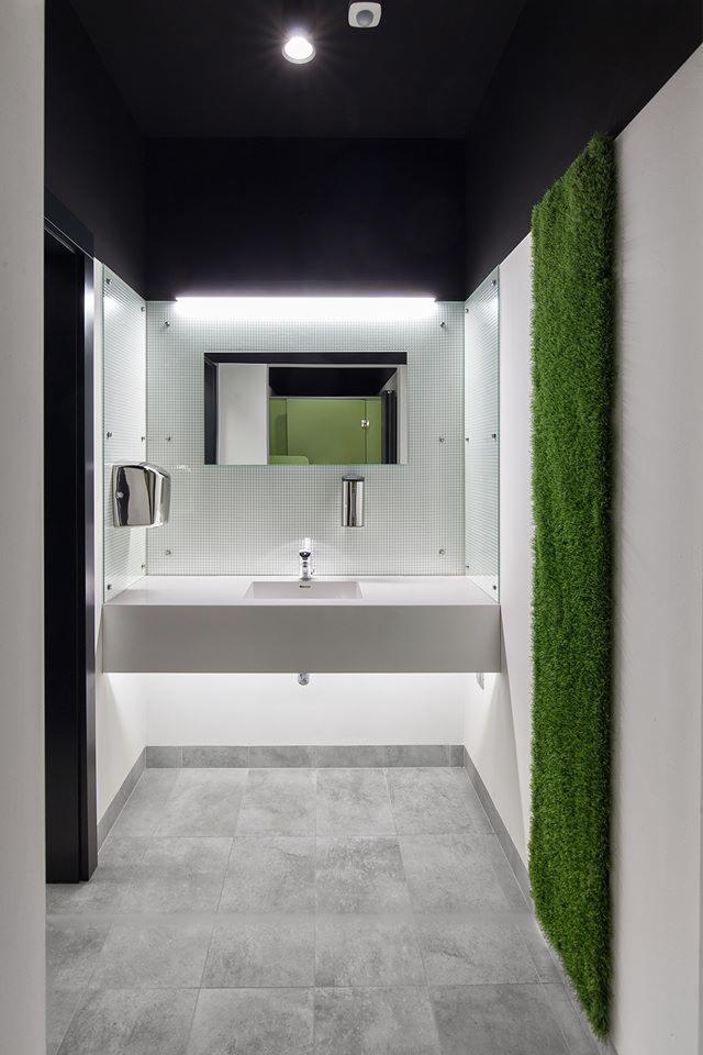 Nowoczesna toaleta publiczna z umywalką nablatową, podświetlanym lustrem na tle mozaiki, pionowym panelem z zielonej trawy i szarymi płytkami podłogowymi.