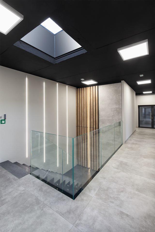 Nowoczesna klatka schodowa z szarą podłogą, szklaną balustradą z czarnymi elementami mocującymi, oświetleniem LED wzdłuż ściany i drewnianą dekoracją ścienną, widok z góry na schody.