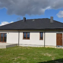 Nowoczesny dom jednorodzinny z ciemną dachówką i jasną elewacją, widok z ogrodu w słoneczny dzień, betonowy taras w trakcie budowy.