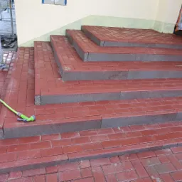 Czerwone, ceglane schody wejściowe po umyciu, z zielonym wężem ogrodowym na pierwszym planie, widoczne rusztowanie i folia ochronna w tle.