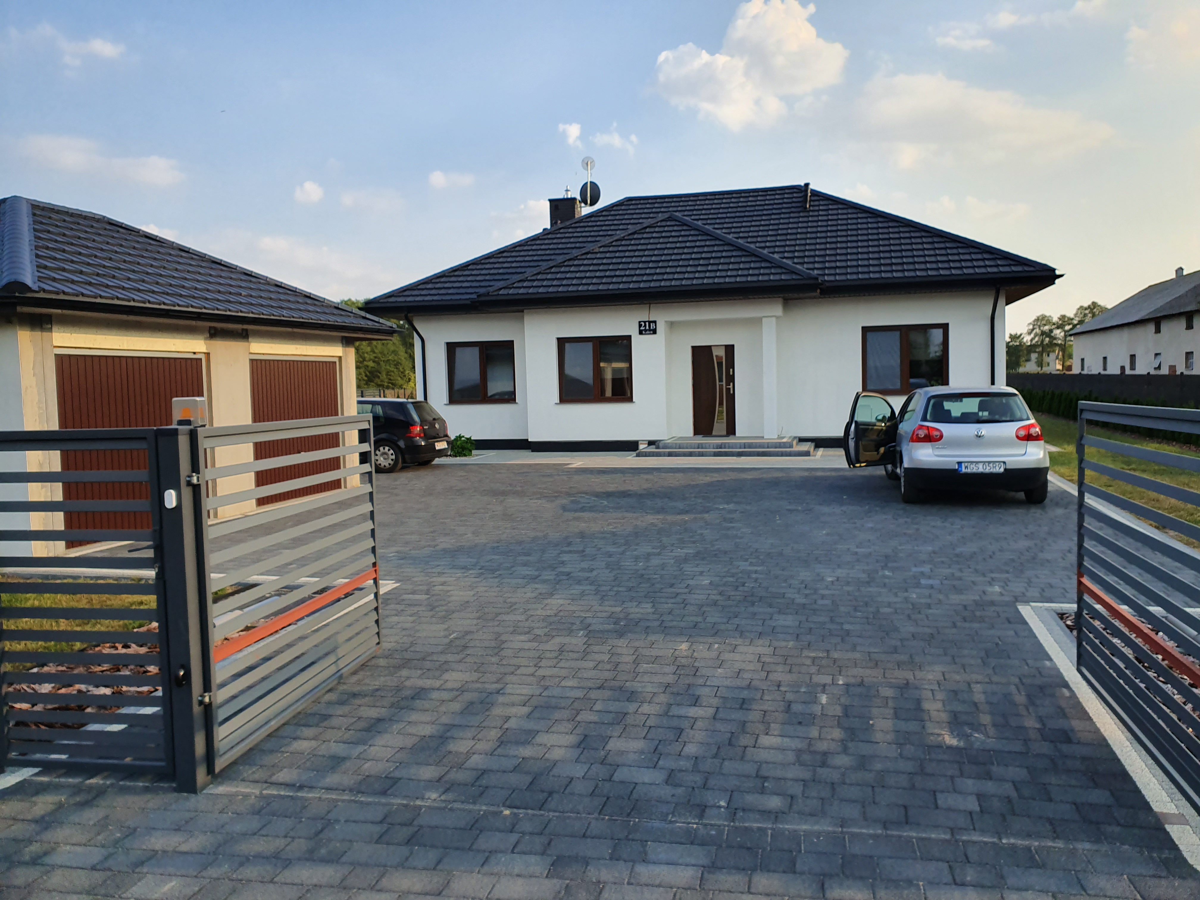 Nowoczesny, parterowy dom z ciemną dachówką, jasną elewacją i dwoma samochodami na posesji, widziany zza szarej, poziomej bramy wjazdowej.