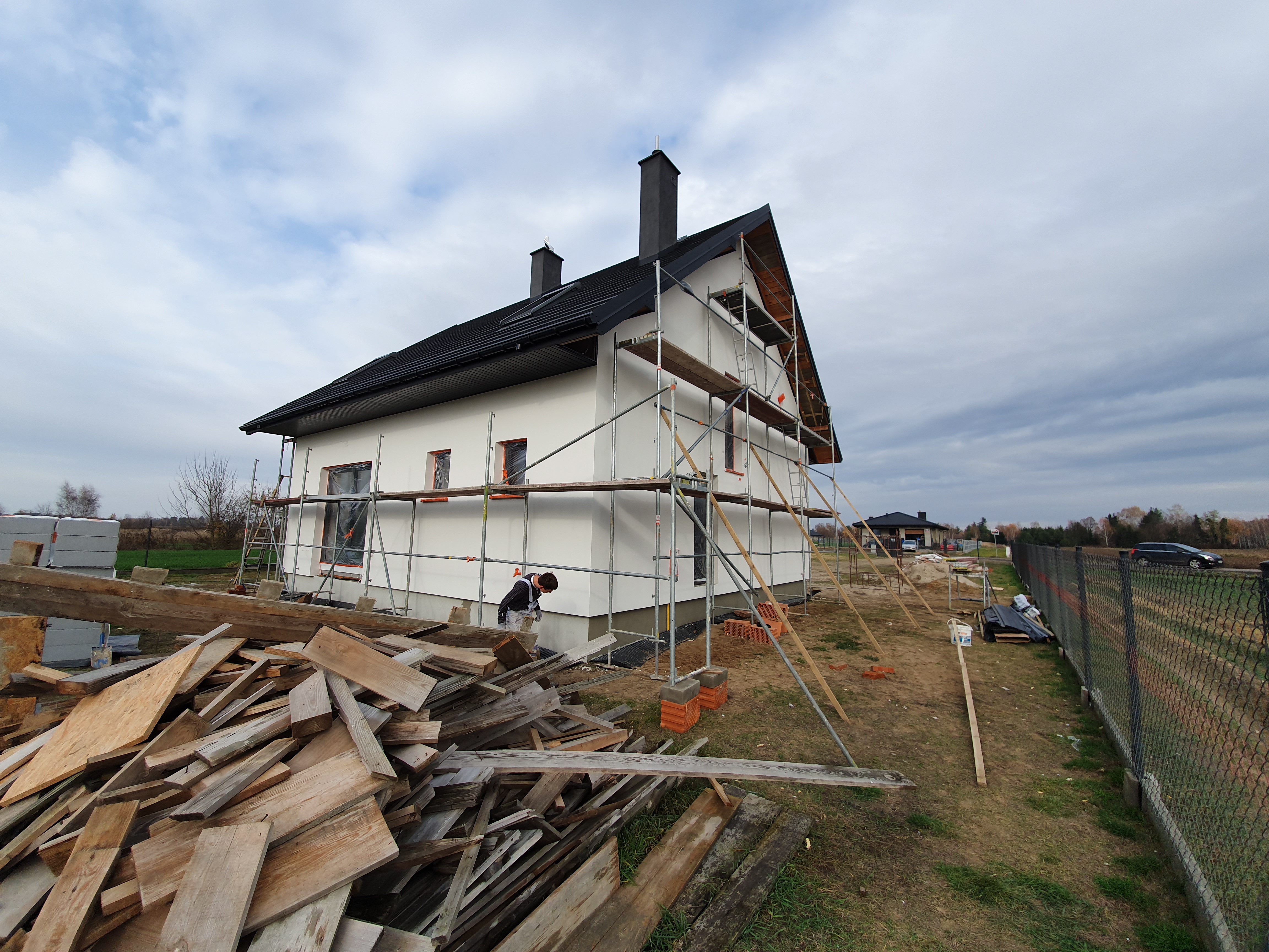 Dom jednorodzinny w trakcie prac elewacyjnych; widoczne rusztowania, stos drewna oraz pracownik w kombinezonie ochronnym przy ścianie