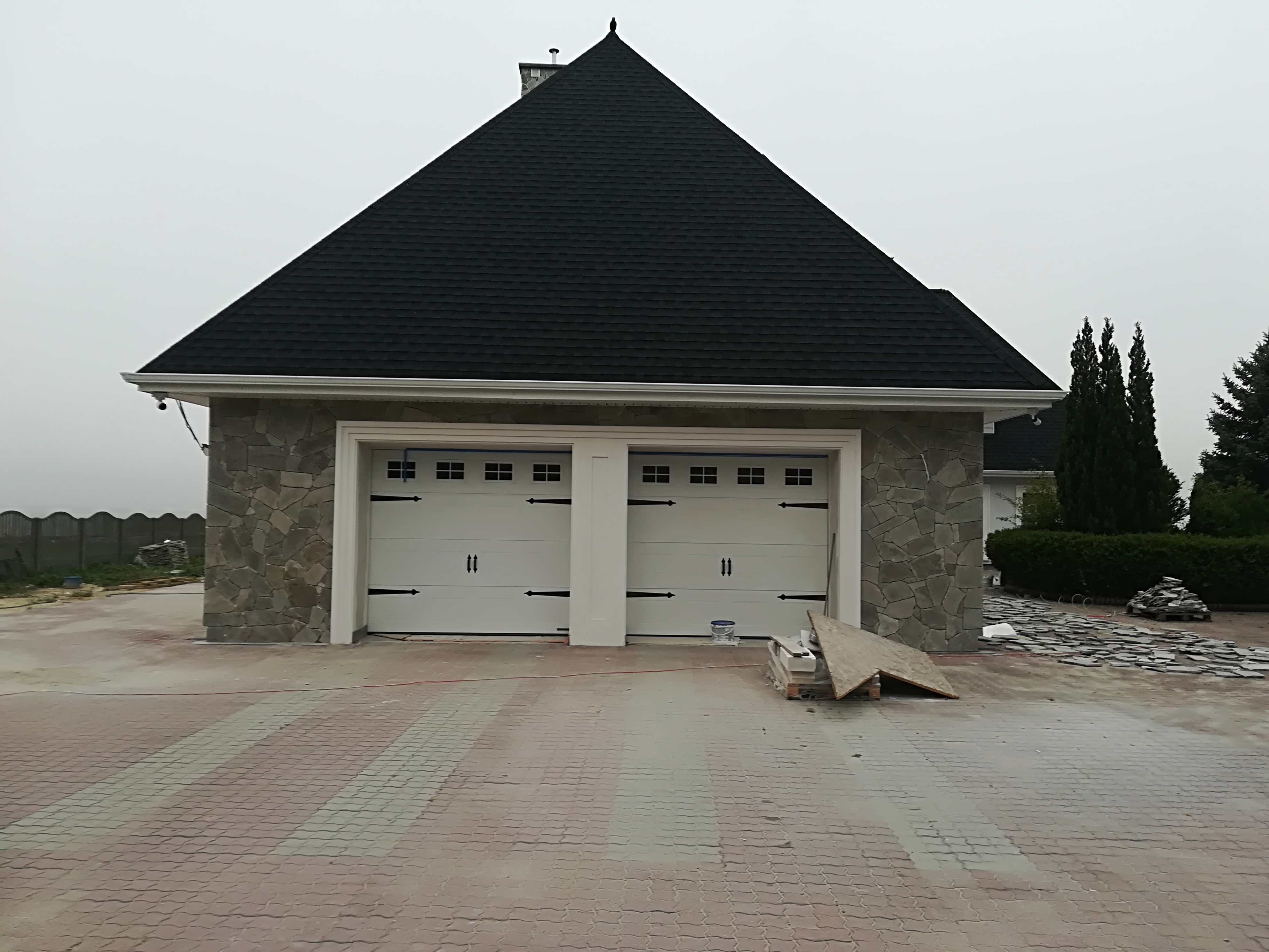 Dwustanowiskowy garaż z białymi drzwiami i elewacją z szarego kamienia, czarny dach z gontu bitumicznego, na podjeździe ułożona kostka, widoczne materiały budowlane i pojemnik z farbą.
