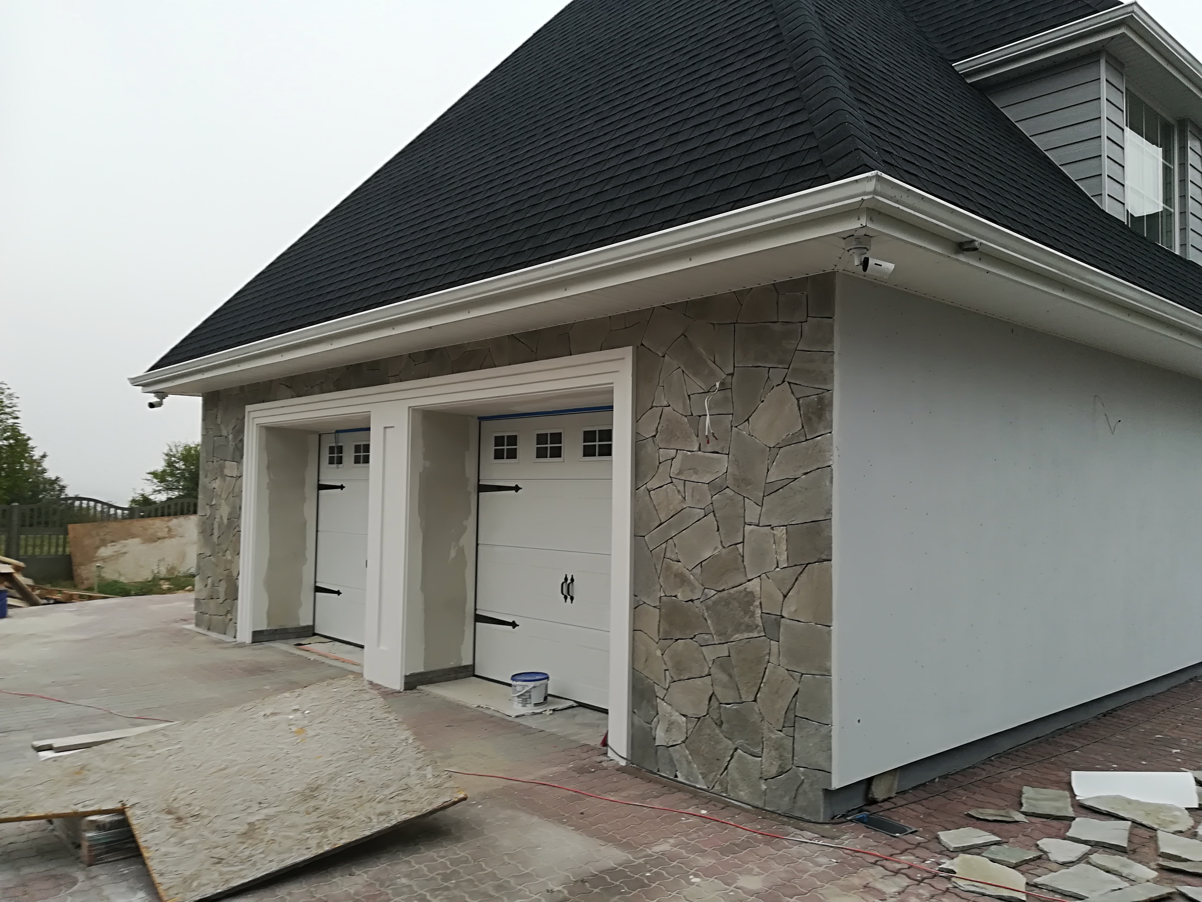 Dwustanowiskowy garaż z elewacją częściowo wykończoną kamieniem naturalnym w trakcie prac, widoczny biały kubeł farby i płyty OSB na podłożu z kostki.