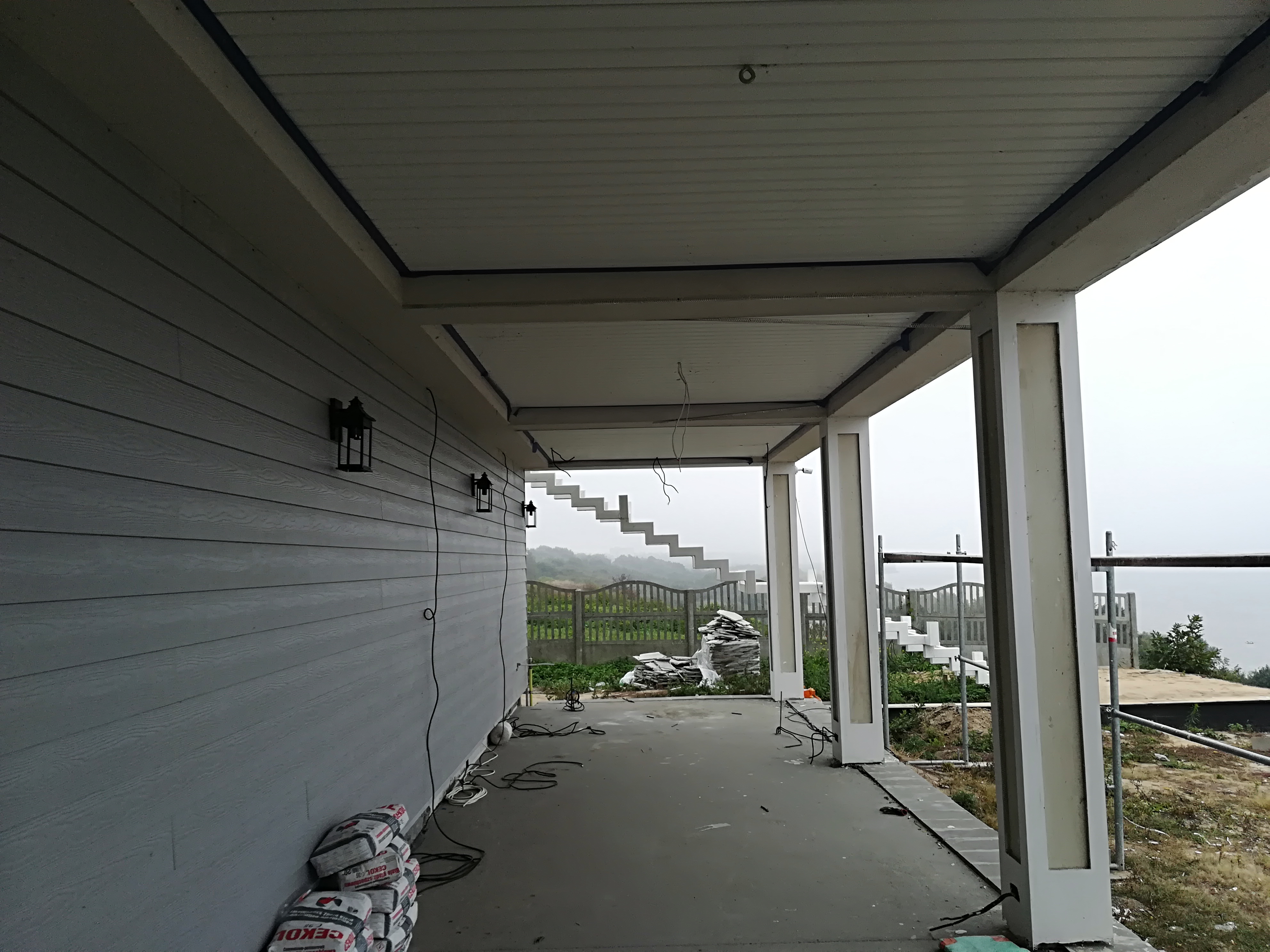 Szara elewacja domu z zamontowanymi czarnymi lampami zewnętrznymi. Widoczne worki z zaprawą murarską, kable elektryczne i elementy konstrukcyjne tarasu w surowym stanie. W tle zarys mglistego...