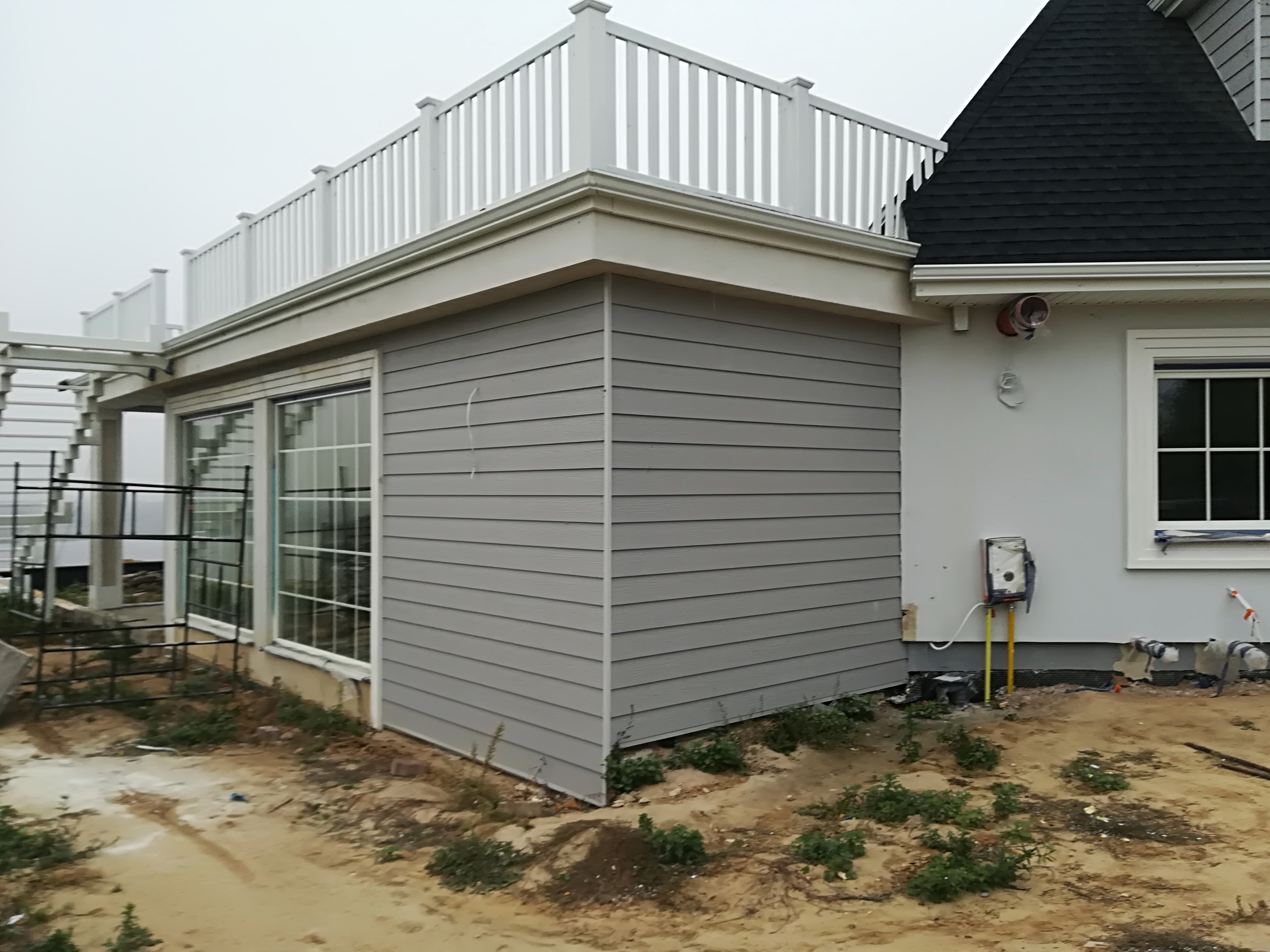 Fragment domu z szarą elewacją sidingową, białymi ramami okiennymi i balustradą na dachu, widoczne elementy instalacji zewnętrznej i teren wokół budynku w trakcie prac wykończeniowych.