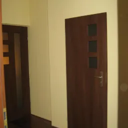 Wnętrze korytarza z dwiema ciemnymi, drewnianymi drzwiami, jedna z trzema małymi szybkami, na tle jasnożółtych ścian i ciemnej, drewnianej podłogi.