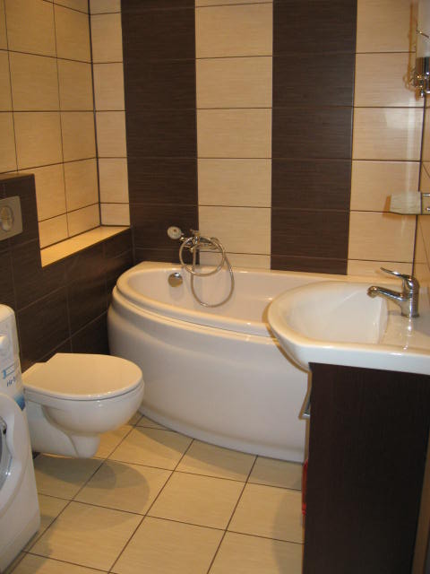 Wykończona łazienka z wanną narożną, umywalką z szafką w kolorze ciemnego brązu i toaletą, wyłożona jasnymi płytkami z ciemnobrązowymi pasami.