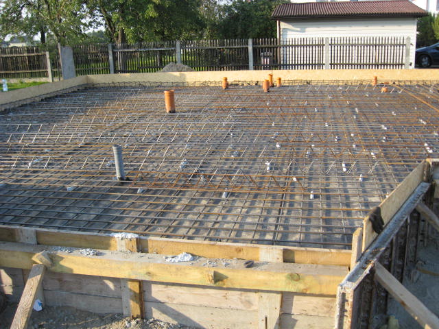 Zbrojenie płyty fundamentowej z rurami kanalizacyjnymi wystającymi pionowo, widoczne drewniane szalunki i otoczenie budowy.