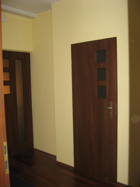 Wnętrze korytarza z dwiema ciemnymi, drewnianymi drzwiami, jedna z trzema małymi szybkami, na tle jasnożółtych ścian i ciemnej, drewnianej podłogi.