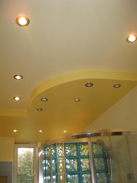 Żółty, podwieszany sufit z wbudowanymi punktowymi lampami LED nad kabiną prysznicową z luksferami.