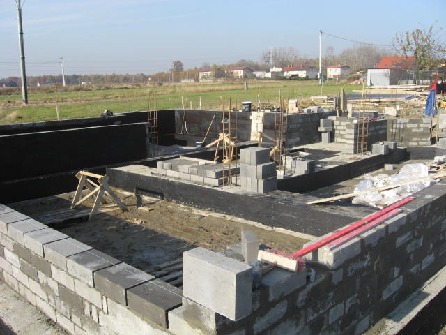 Wykopane ławy fundamentowe z izolacją bitumiczną i częściowo wymurowanymi ścianami z bloczków betonowych, widoczne zbrojenia, drewniane stemple i pejzaż wiejski w tle.