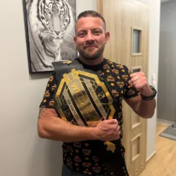 Uśmiechnięty mężczyzna z brodą, trzymający pas mistrzowski WKDZIK na tle ściany z obrazem tygrysa. Ubrany w koszulkę z wzorem MCM i jeansy.