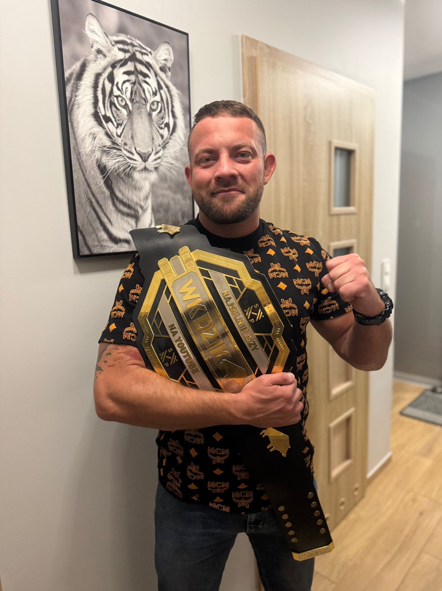 Uśmiechnięty mężczyzna z brodą, trzymający pas mistrzowski WKDZIK na tle ściany z obrazem tygrysa. Ubrany w koszulkę z wzorem MCM i jeansy.