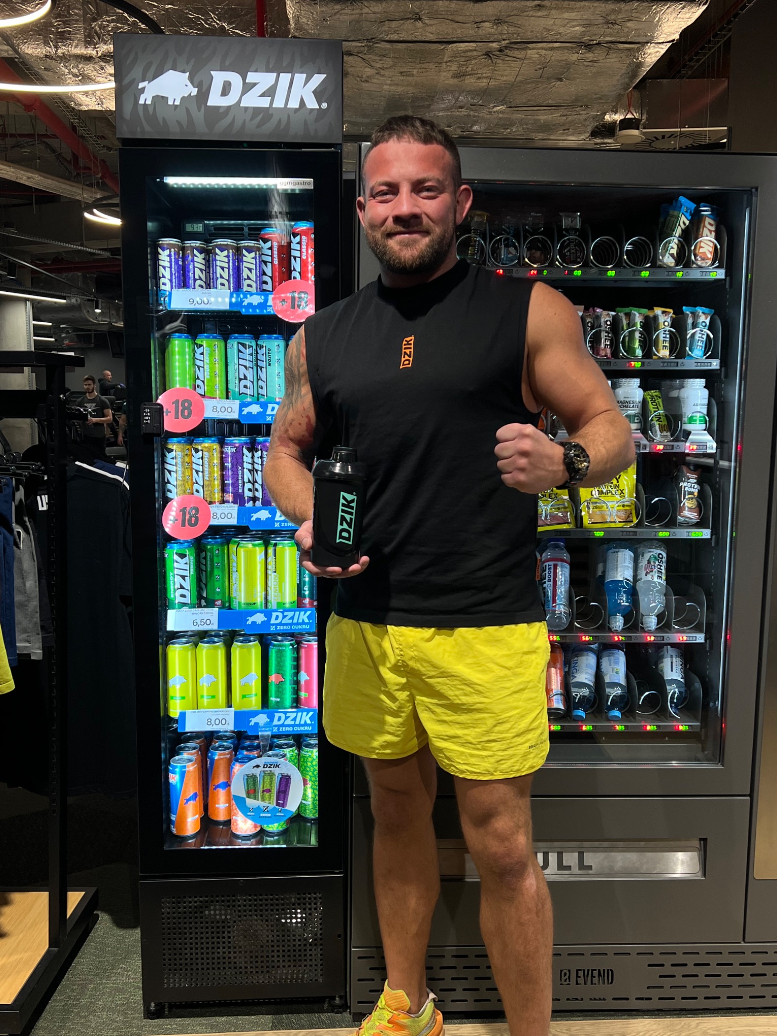Mężczyzna w sportowym stroju pozuje w Szczecinie obok automatu z napojami energetycznymi marki Dzik, trzymając shaker. Widoczny tatuaż na ramieniu.