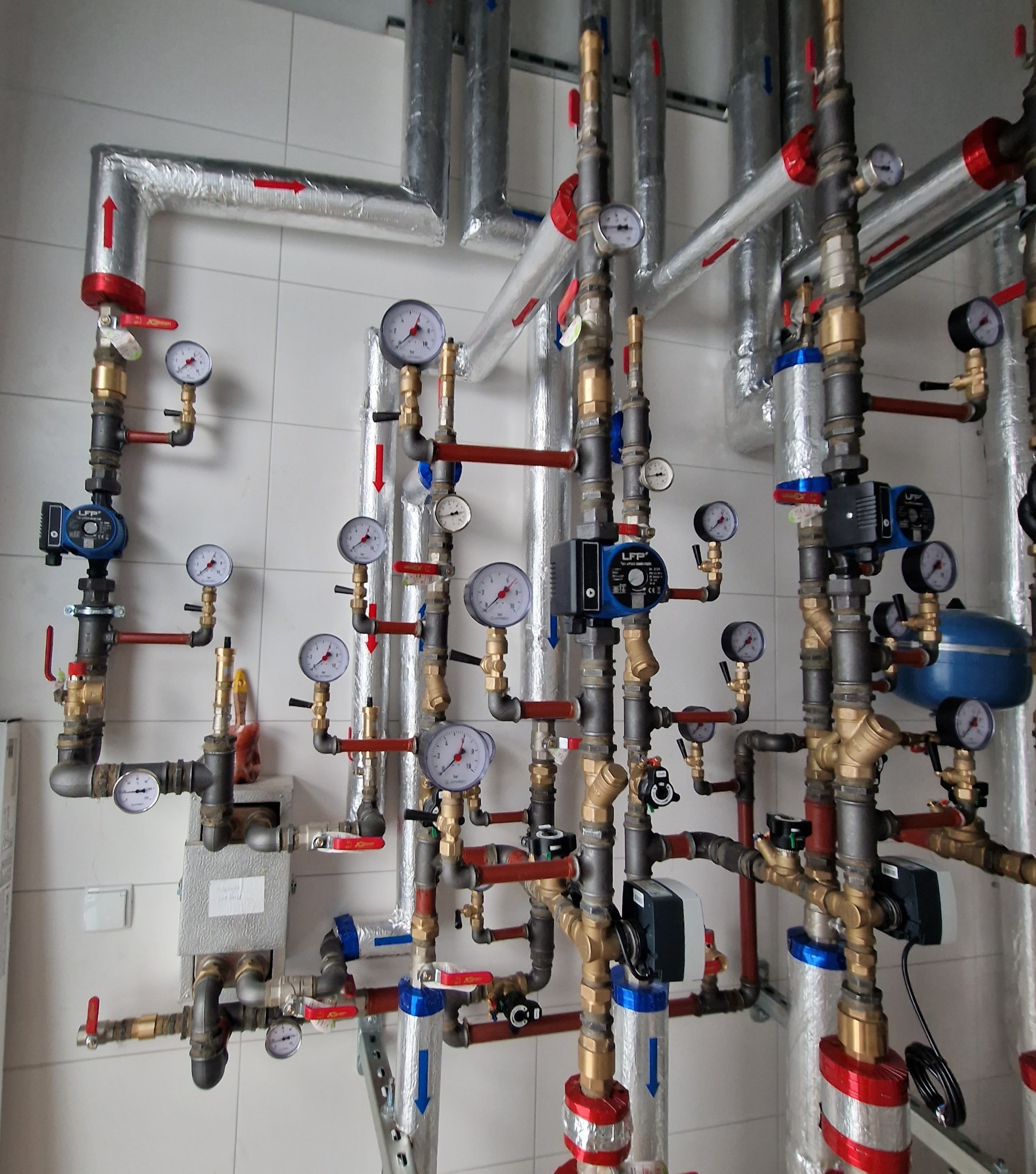 Złożona instalacja rur i zaworów z manometrami na białej, kafelkowanej ścianie. Rury są izolowane folią z widocznymi strzałkami kierunku przepływu. Widoczne pompy obiegowe i filtry.