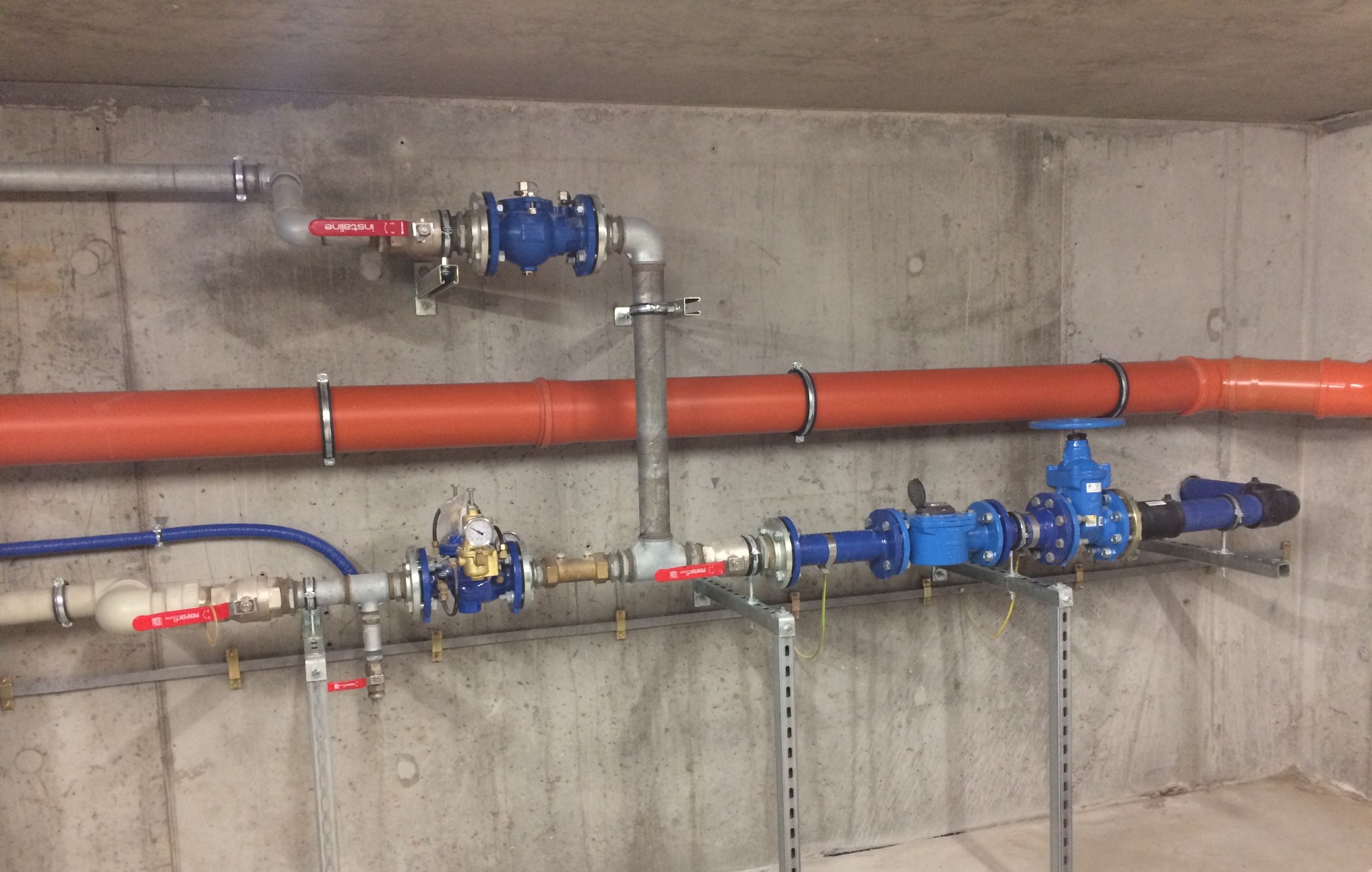 Różnokolorowe rury i zawory instalacji wodnej zamontowane na betonowej ścianie wewnątrz budynku, widoczne elementy mocujące i manometr.