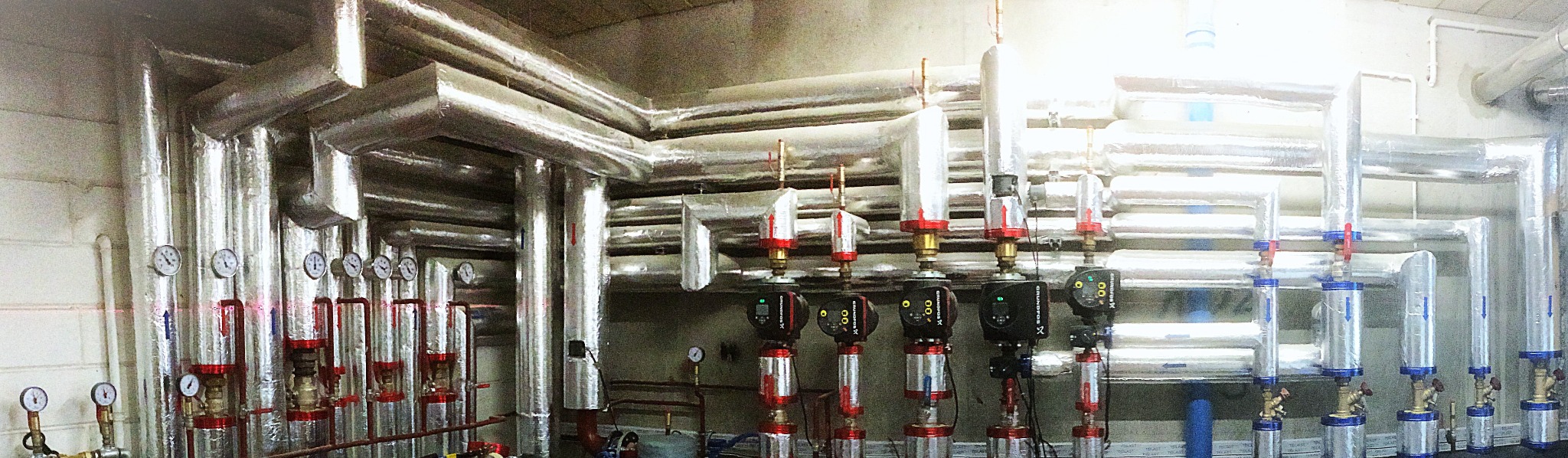 Rozbudowana instalacja grzewcza z izolowanymi rurami, pompami obiegowymi Grundfos i manometrami, widok na kotłownię.