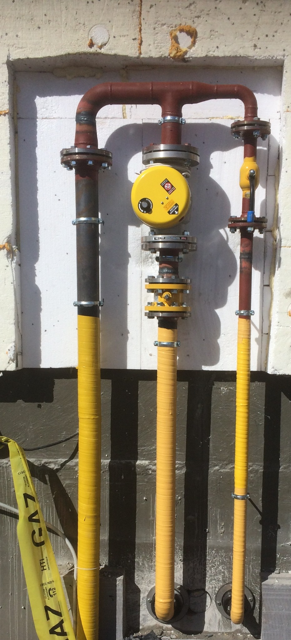Zewnętrzna instalacja gazowa z żółtymi rurami, widoczne elementy przyłączeniowe i licznik gazu, na tle ocieplonej ściany budynku z widoczną taśmą ostrzegawczą GAZ.