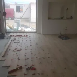 Układanie paneli podłogowych w nowym domu; widoczne poziomice, kliny dystansowe i pracownik przy oknie.