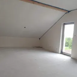 Surowe wnętrze poddasza z oknami i przygotowanymi otworami na gniazdka, ściany i sufit pokryte świeżym tynkiem, widoczne drewniane elementy konstrukcyjne pod sufitem.