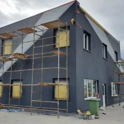 Dwukondygnacyjny budynek w trakcie malowania elewacji na szaro z białymi pasami, osłonięte okna, rusztowania, puszki z farbą i zaparkowany samochód na zewnątrz.