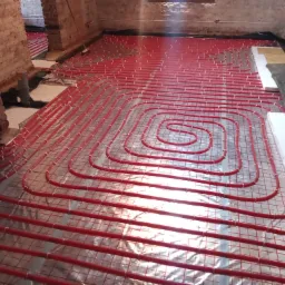 Układanie czerwonych rur ogrzewania podłogowego na folii odblaskowej w pomieszczeniu z ceglanymi ścianami, widoczne elementy izolacji