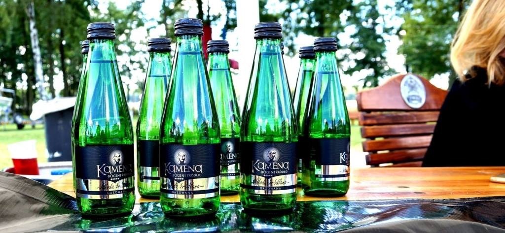 Szklane butelki wody gazowanej Kamena z czarnymi etykietami ustawione na drewnianym stole, w tle park z zielenią i ławką.