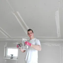 Uśmiechnięty mężczyzna w ubraniu roboczym, trzymający w rękawicach szpachelki z nałożoną masą szpachlową, na tle pomieszczenia z płytami kartonowo-gipsowymi i częściowo wyszpachlowanym sufitem.