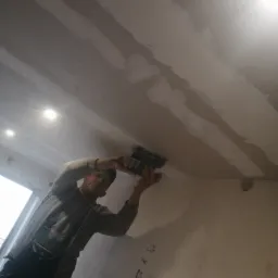 Pracownik w kombinezonie szlifuje szpachlowany sufit w Tarnawatce za pomocą szlifierki elektrycznej, widoczne ślady po szpachlowaniu i notatki na ścianie.