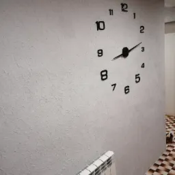 Ściana z nałożoną fakturą, na której zamontowano duży, czarny zegar ścienny z minimalistycznymi cyframi, widoczny fragment grzejnika i fragment podłogi w kratkę w tle.