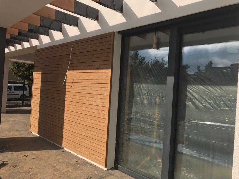 Nowoczesna elewacja budynku z poziomymi panelami imitującymi drewno, widoczne przesuwne okna z ciemnymi ramami i fragment pergoli z czarną folią ochronną na dachu.