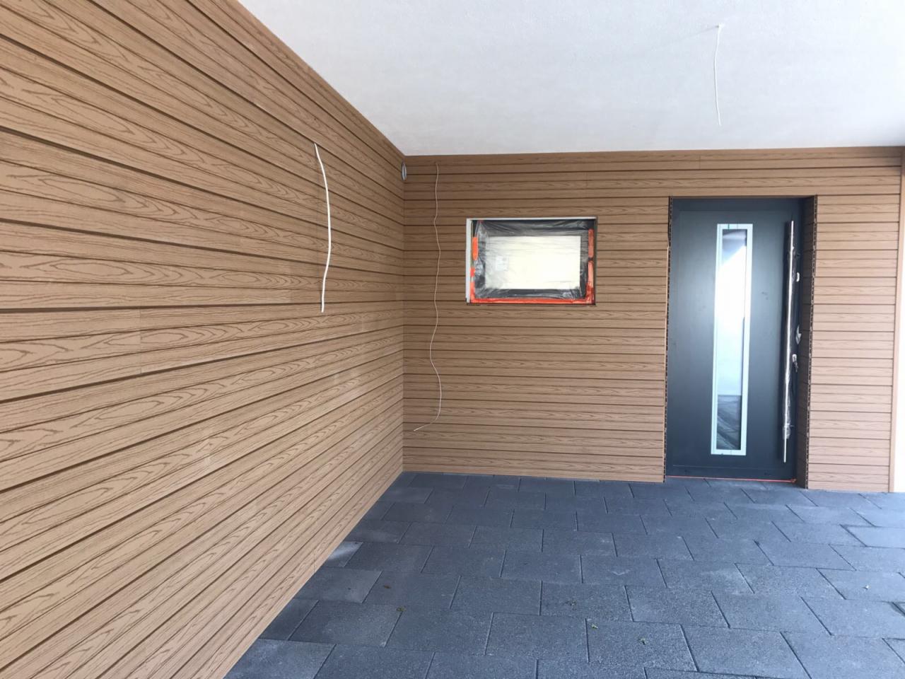 Zadaszony taras z wykończeniem ścian panelami imitującymi drewno, z widocznym oknem zabezpieczonym folią i nowoczesnymi drzwiami wejściowymi.