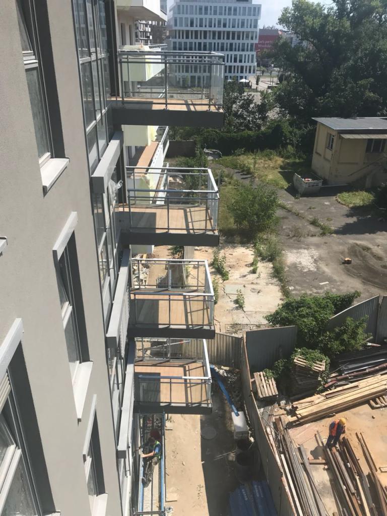 Wysokie budynki z balkonami z metalowymi balustradami i szklanymi panelami, widok z góry na plac budowy z robotnikami i materiałami budowlanymi.