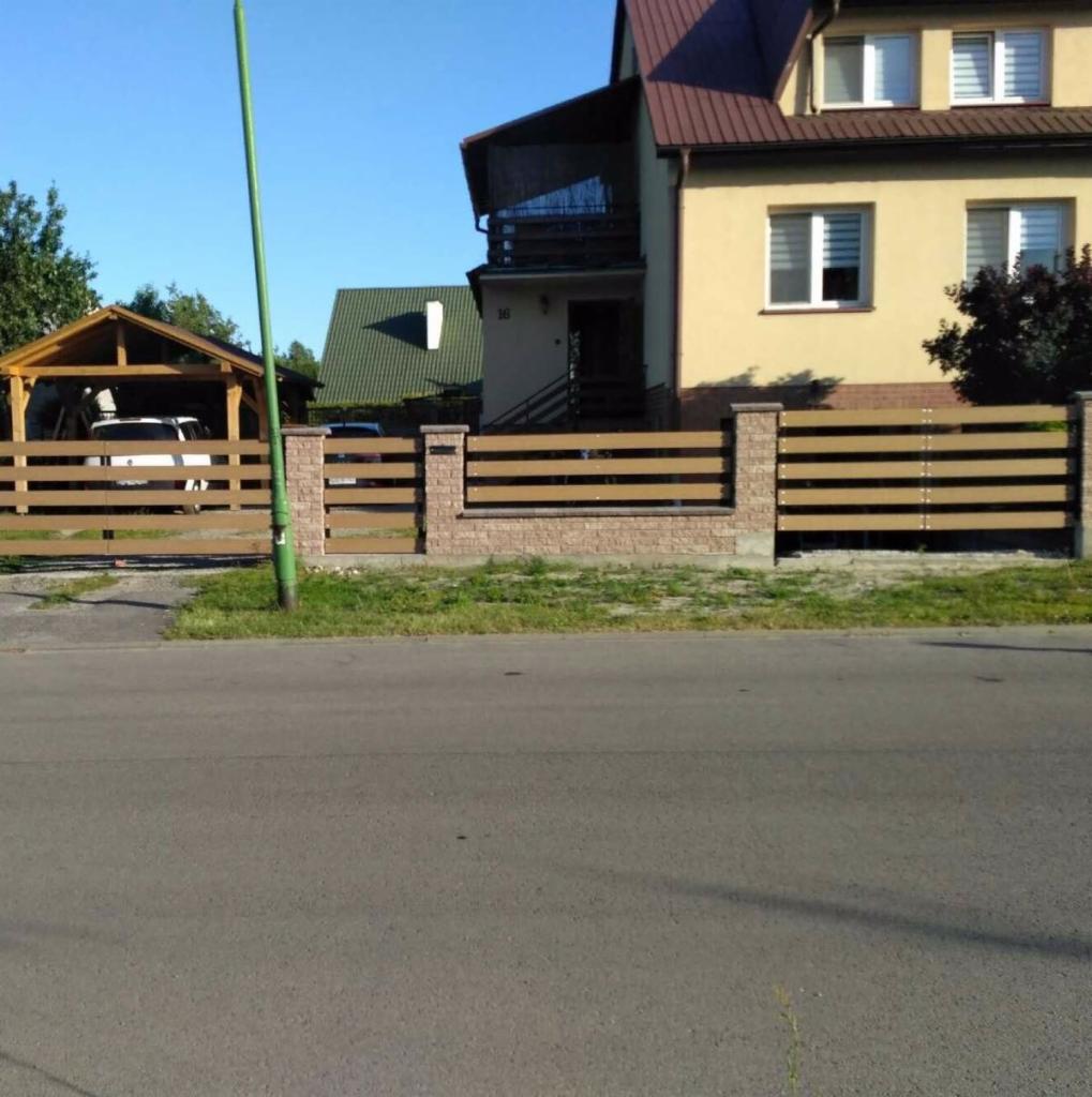 Nowoczesne ogrodzenie z poziomych, brązowych desek i kamiennych słupków przed domem z balkonem i wiatą garażową.