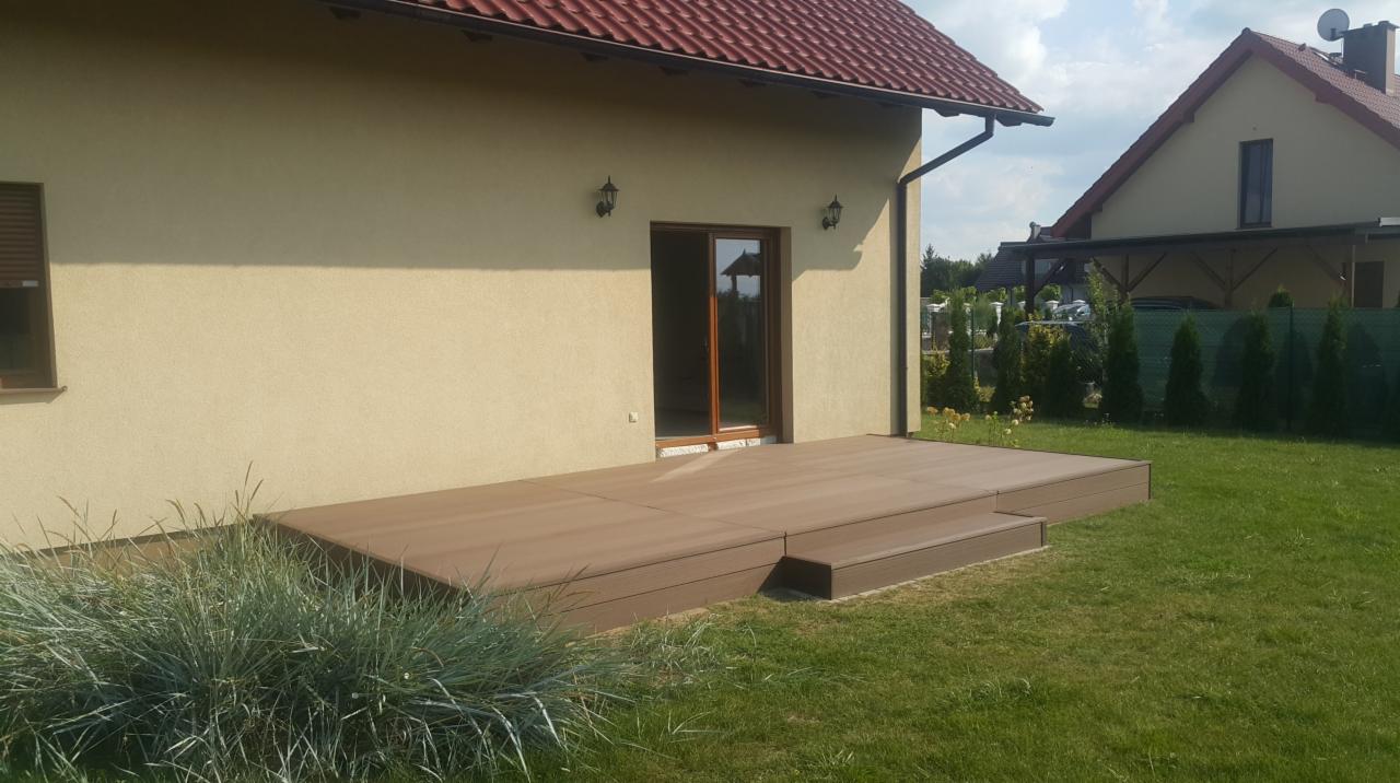 Brązowy taras kompozytowy z jednym stopniem, przylegający do beżowej ściany domu z brązowymi drzwiami balkonowymi, otoczony zielonym trawnikiem i ozdobną trawą.