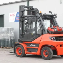 Pomarańczowy wózek widłowy Linde na zewnątrz budynku magazynowego z częściowo otwartą bramą i paletą z zapakowanymi pojemnikami.