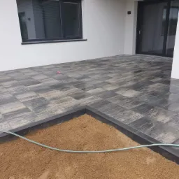 Częściowo ułożony taras z szarej kostki z widocznym piaskowym podłożem i wężykiem ogrodowym, w tle biała ściana budynku z oknami i przesuwnymi drzwiami.