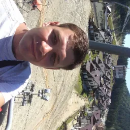 Autoportret z wyciągu krzesełkowego, w tle budowa osiedla domków jednorodzinnych z ciemnymi dachami i budynek w trakcie budowy z dźwigiem.