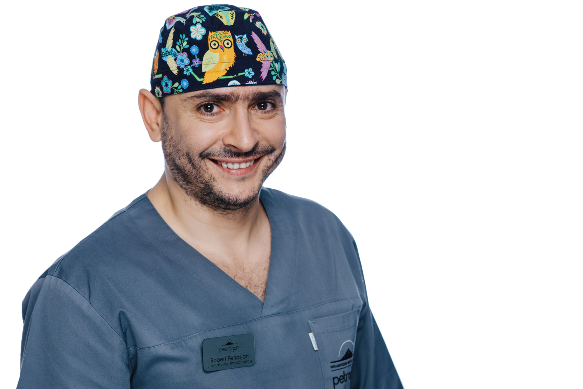 Uśmiechnięty stomatolog Robert Petrosjan w czepku z motywem sów i kwiatów, ubrany w szary uniform z identyfikatorem, na białym tle.
