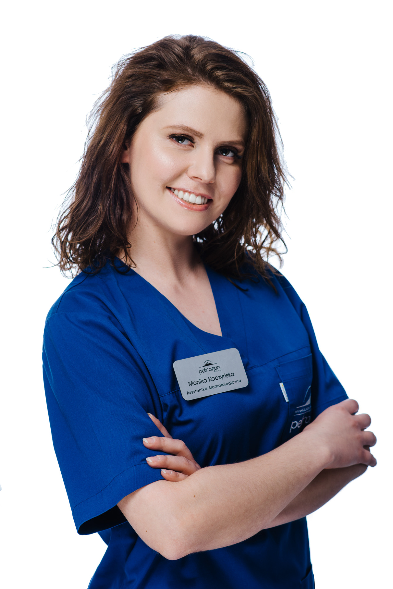 Portret uśmiechniętej asystentki stomatologicznej Moniki Kaczyńskiej w niebieskim uniformie z identyfikatorem 'Petrosian Clinic', skrzyżowane ręce na białym tle.