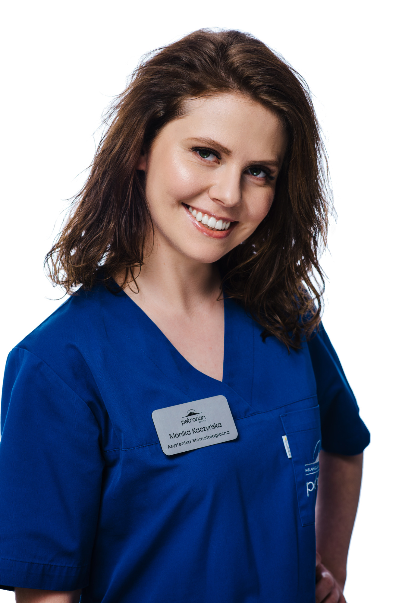 Uśmiechnięta asystentka stomatologiczna Monika Kaczyńska w niebieskim uniformie, z identyfikatorem kliniki Petrasian Clinic, pozuje na białym tle.