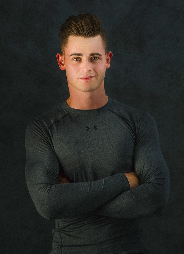 Młody mężczyzna w szarej koszulce termoaktywnej z długim rękawem, pozujący na ciemnym, teksturowanym tle z założonymi rękami.