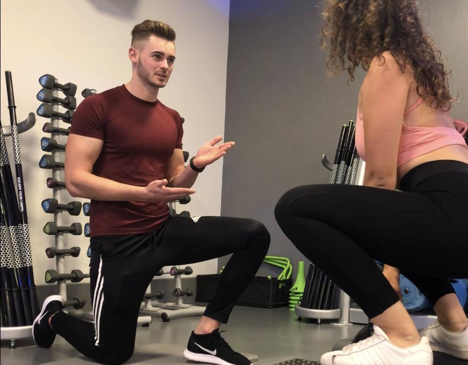 Instruktor fitness na jednym kolanie demonstruje ćwiczenie kobiecie w sportowym stroju, w tle stojak z hantlami i sprzęt do ćwiczeń.