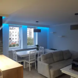 Jasne, nowocześnie urządzone wnętrze salonu z jadalnią, z oknami podświetlonymi niebieskim światłem LED, szarym narożnikiem i stołem z krzesłami.