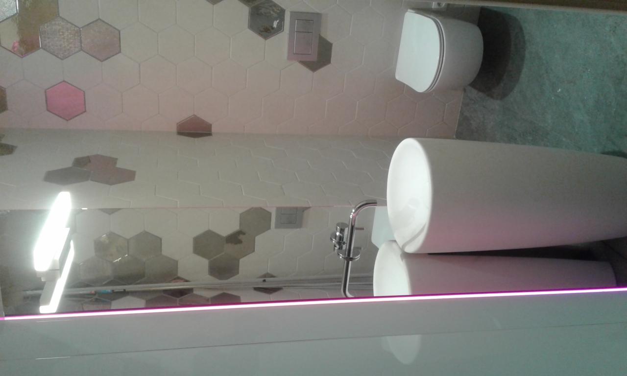 Nowoczesna łazienka z heksagonalnymi płytkami w odcieniach bieli, szarości i różu, wisząca toaleta i umywalka nablatowa z baterią, odbicie w lustrze z podświetleniem LED.