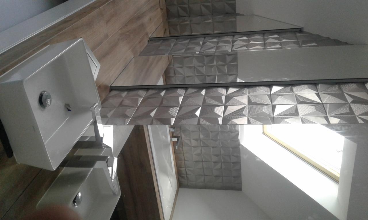 Ukończona łazienka z umywalką i ścianą wyłożoną szarymi, trójwymiarowymi płytkami ceramicznymi o geometrycznym wzorze, oświetlona światłem z okna dachowego.